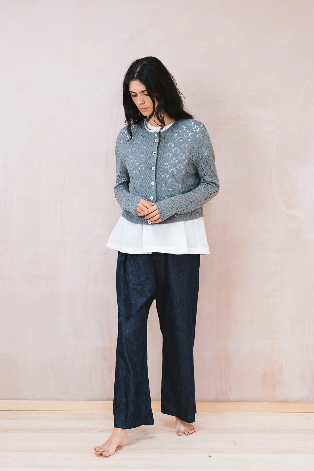 Person wearing a gray cardigan, white top, and dark blue pants against a plain background